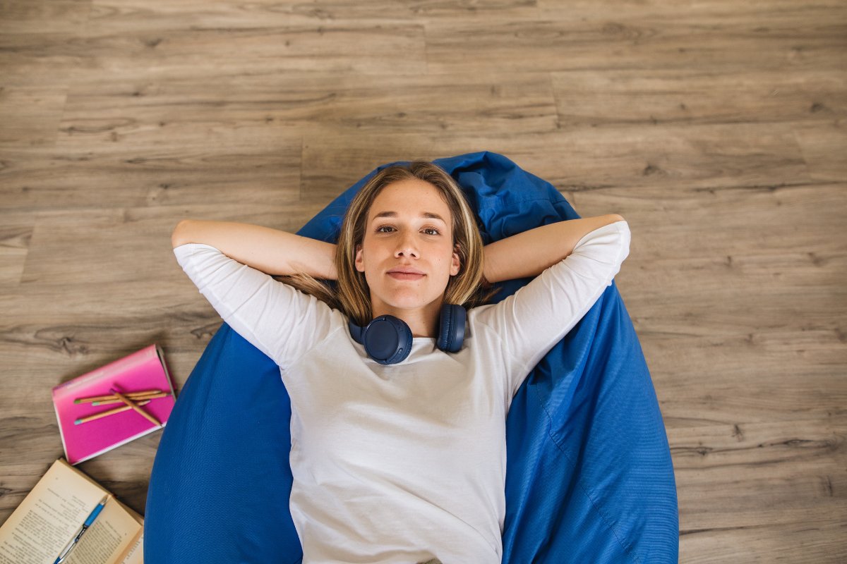 Étudiante allongée sur un pouf, casque audio autour du cou, à côté de ses cahiers et d'un livre ouvert